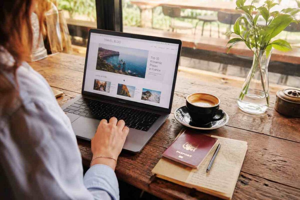 Woman browsing latest articles TravellingApples travel blog on a laptop with passport, notebook, and coffee on a wooden café table