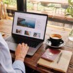 Woman browsing latest articles TravellingApples travel blog on a laptop with passport, notebook, and coffee on a wooden café table